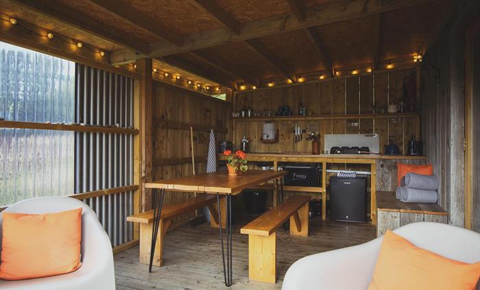 A camp kitchen with festoon lighting above, a countertop with shelving above with glasses and mugs on. A fridge is below the counter and in the foregr